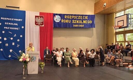 Zdjęcie z inauguracji roku szkolnego 2026/2027 w sali gimnastycznej. Kobieta przemawia za mównicą na tle dekoracji z napisem "ROZPOCZĘCIE ROKU SZKOLNEGO 2026/2027" oraz godłem Polski. Widoczna publiczność siedząca na krzesłach i ławkach.